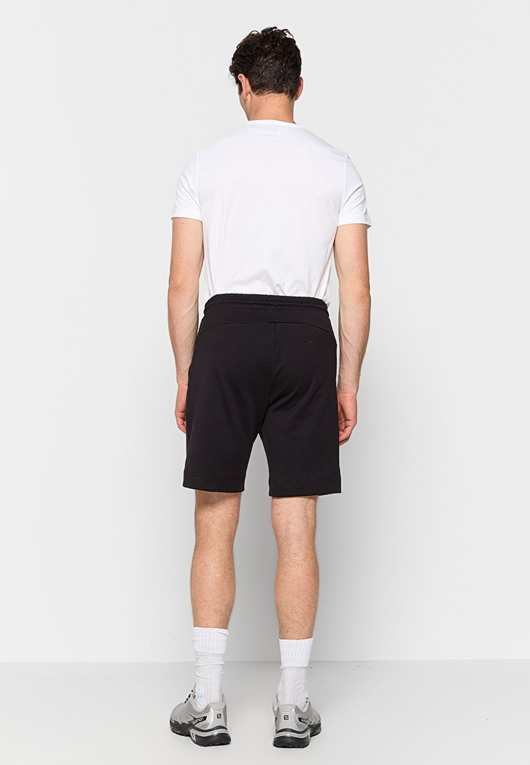 Czarne spodenki z elastycznym paskiem, wykonane z miękkiego materiału, połączone z prostą białą koszulką i szarymi butami sportowymi.