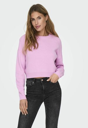 Pull court rose clair avec un col rond, des poignets côtelés et une texture lisse, associé à un jean noir.