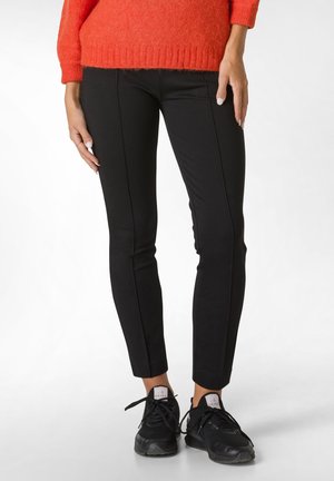 Schwarze, figurbetonte Hosen mit glatter Textur und seitlichen Nähten, kombiniert mit schwarzen Sportschuhen. Auch ein leuchtend roter Pullover ist sichtbar.