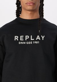 Černá mikina s kulatým výstřihem a dlouhými rukávy, na přední straně s textem "REPLAY DNM GDS 1981" v stříbrné výšivce.