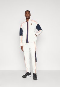 Λευκή φόρμα με ναυτικό μπλε λεπτομέρειες και πορτοκαλί ραφές. Ζακέτα με φερμουάρ και αντίστοιχες παντελόνες. Μαύρα αθλητικά παπούτσια.