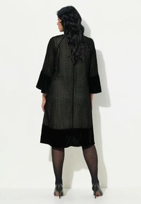 Schwarzes Kleid aus strukturiertem Stoff mit einem Karomuster, Glockenärmeln und Samt-Details am Saum. Kombiniert mit transparenten Strumpfhosen und hohen Schuhen.