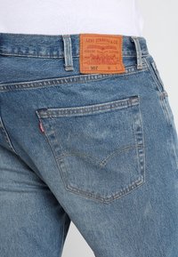 Jeans in denim blu con una toppa in pelle marrone chiaro sulla vita posteriore, caratterizzati dal logo di Levi Strauss e un classico design a cinque tasche.