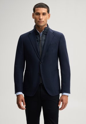 Mann trägt einen dunkelblauen Blazer über einer schwarzen Reißverschlussjacke und einem hellblauen Hemd, steht vor einem einfarbigen hellgrauen Hintergrund.