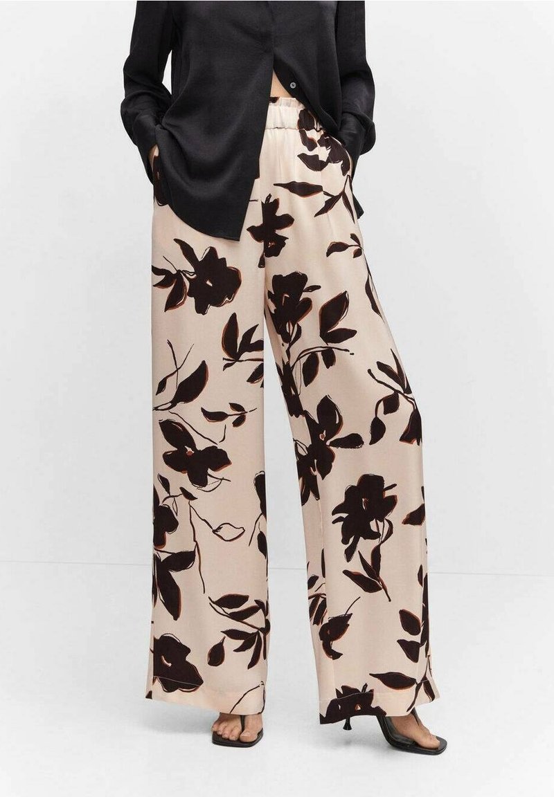Pantalon large beige à imprimé floral noir, ceinture élastique et tissu doux. Associé à une chemise noire et des sandales plates.