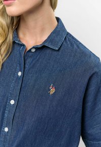 Tmavomodrá riflová košeľa s golierom, s bielymi gombíkmi a malým vyšitým logom na ľavej hrudi. Hladká textúra.