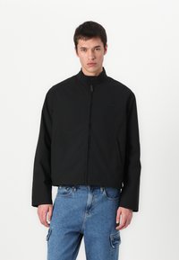 Jachetă neagră scurtă cu guler în picioare, închidere cu fermoar și buzunare laterale. Fabricată dintr-un material neted, cu un design minimalist.