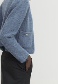 Pull en laine léger et flou de couleur bleu clair, avec une poche latérale et un détail de bouton doré, associé à un pantalon noir ajusté.