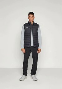 Black puffer vest, lightweight and quilted, worn over a gray long-sleeve shirt. Model in black pants and gray athletic shoes. Simple design.