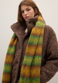 Manteau en polaire marron avec un col haut, en tissu texturé, associé à une écharpe rayée multicolore aux tons verts, jaunes et bruns.
