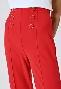 Pantalons rouges à la texture lisse, avec des accents en anneaux dorés et une coupe ajustée. Les coutures verticales ajoutent de la structure au design.