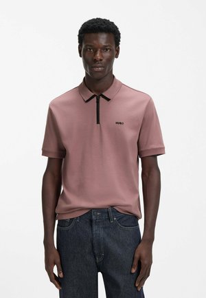 Short-sleeve polo shirt in soft pink fabric, featuring a black zip collar and a small black logo on the chest, paired with dark denim jeans.
