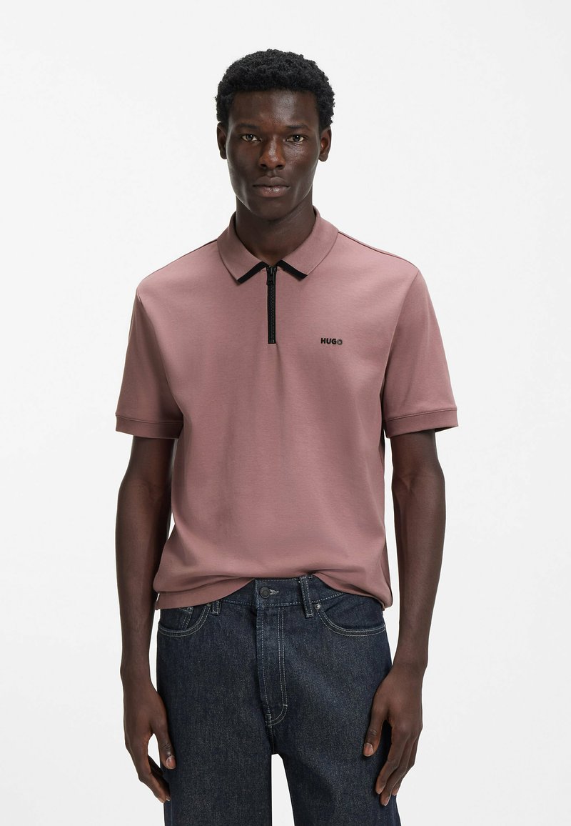 Polo a maniche corte in tessuto rosa tenue, con colletto a zip nero e piccolo logo nero sul petto, abbinata a jeans denim scuro.