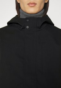 Black hooded jacket with a zip closure, featuring a high collar and a subtle texture. Includes snap buttons and a small side pocket.