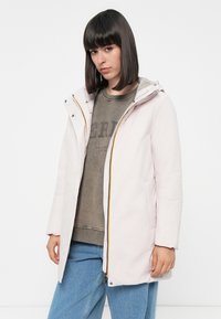 Chaqueta con capucha de color rosa claro y cremallera con detalles en amarillo. Llevada sobre un suéter verde oliva y jeans azules. Textura suave, forma ajustada.
