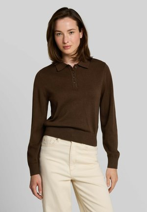 Vrouw met schouderlang bruin haar, gekleed in een bruine polo met lange mouwen en crèmekleurige broek tegen een effen grijze achtergrond.