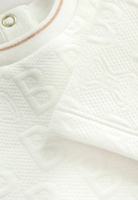 Primer plano de una tela blanca texturizada con sutiles patrones geométricos y un detalle de botón cerca del borde del cuello.