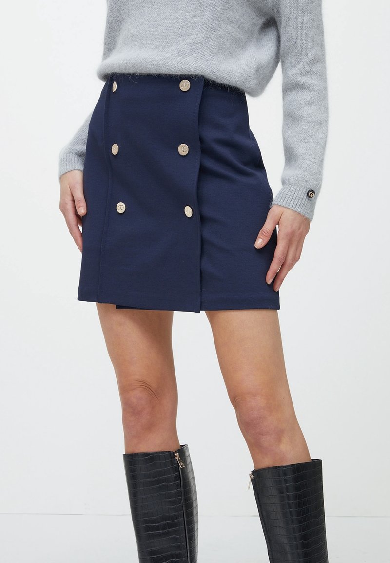 Jupe bleu marine avec un design croisé, ornée de boutons dorés. Le tissu semble lisse et ajusté, associé à des bottes noires montantes.