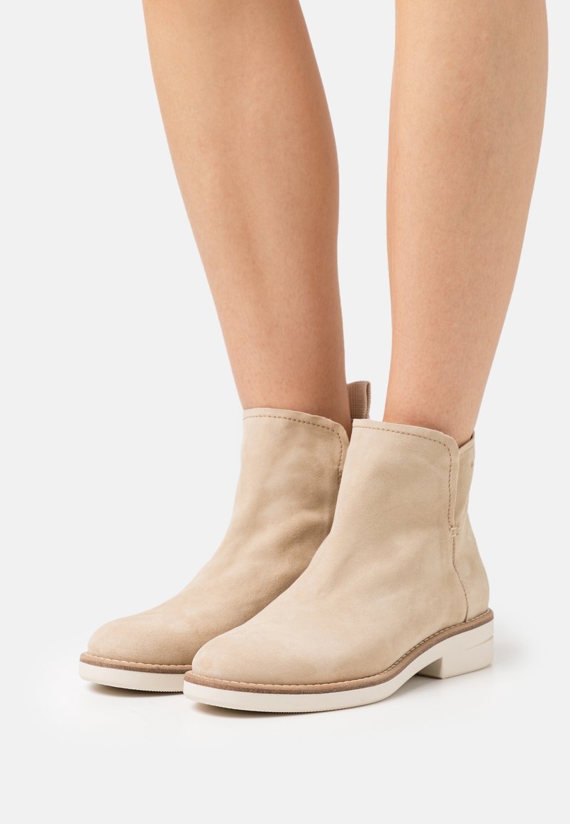 Bottines en daim beige avec languettes, portées sur des jambes nues, dotées de semelles blanches plates et de coutures simples.