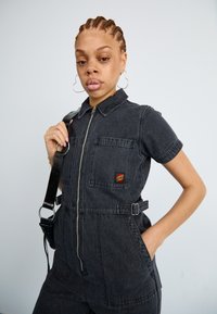 Svart denim jumpsuit med korte ermer, frontglidelås, to lommer og beltehempe. Har en logopatch på brystet.
