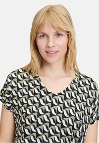 Schwarzes und olivfarbenes gemustertes Oberteil mit V-Ausschnitt und kurzen Ärmeln, mit einem textilem Design aus ineinandergreifenden Formen.