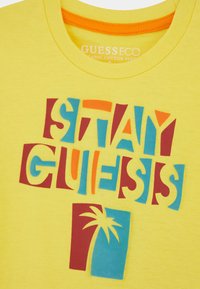 Keltainen luomupuuvillainen t-paita, jossa on rohkeaa moniväristä tekstiä "STAY GUESS" ja palmu-graafinen kuvasto, sekä pyöreä kaula-aukko.