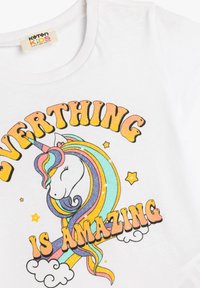 Vit bomulls T-shirt med en färgglad enhörning som har regnbågsfärgat hår och glittrande text som säger "ALLT ÄR AMAZING."