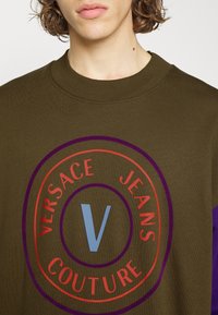 Olivgrön sweatshirt med ribbade mudd. Har en cirkulär logotyp i röd, lila och blå text: "VERSACE JEANS COUTURE" och en central 'V'.