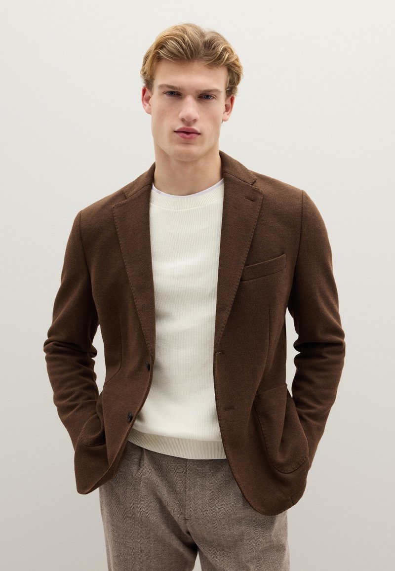 Braunes Sakko mit strukturierter Oberfläche, einer Brusttasche und zwei unteren Taschen, kombiniert mit einem weißen, gerippten Pullover und hellbraunen Hosen.