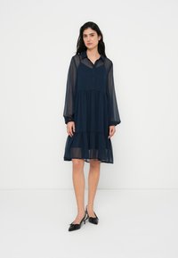 Φόρεμα σε ναυτικό μπλε χρώμα, διαφανές, με μακριά μανίκια, εμπρόσθιο κλείσιμο με κουμπιά και φούστα με στρώσεις. Διαθέτει κλασικό γιακά και απαλό υφή. Μαύρα παπούτσια με φιόγκους.