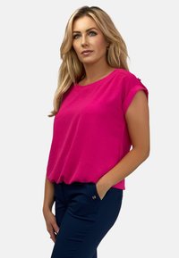 Krótki rękaw, jasnoróżowa bluzka o luźnym kroju i podwiniętych rękawach, zestawiona z granatowymi spodniami z małym dekoracyjnym akcentem.