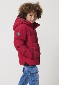 Giacca puffa rossa con cappuccio, caratterizzata da accenti neri e logo sulla manica. Il materiale è liscio, con una texture trapuntata.