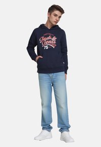 Giovane uomo in piedi con le mani in tasca, indossando un felpa blu scuro con testo grafico, jeans azzurri chiari e sneaker bianche su sfondo neutro.