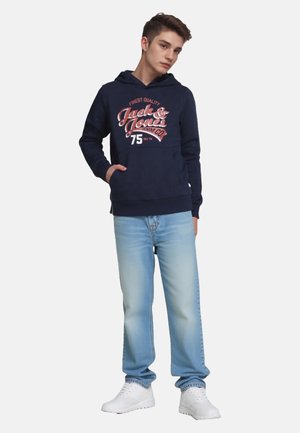 Young man standing with hand in pocket wearing navy hoodie with graphic text, light blue jeans, and white sneakers on plain background.