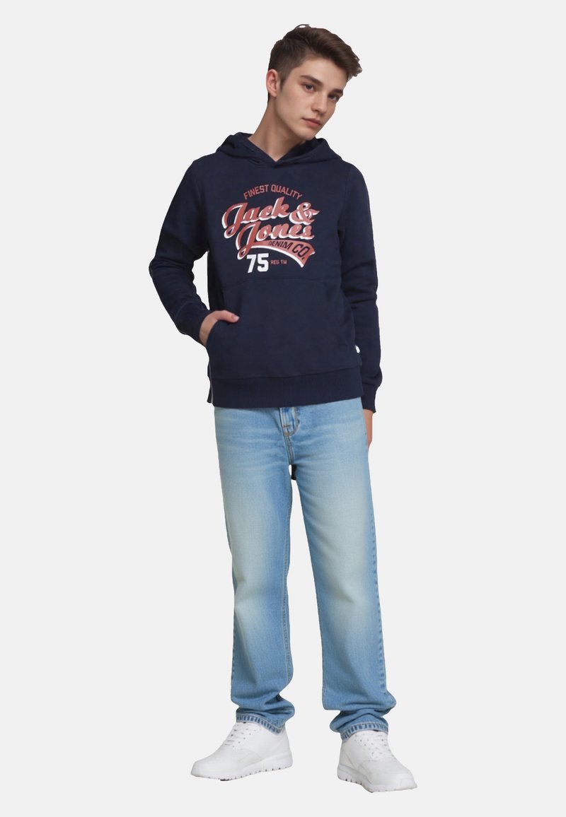 Giovane uomo in piedi con le mani in tasca, indossando un felpa blu scuro con testo grafico, jeans azzurri chiari e sneaker bianche su sfondo neutro.