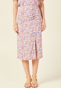 Blumiger Midi-Rock mit einem bunten Druck auf pinkem Hintergrund, mit einem Frontschlitz und einer lockeren, fließenden Form. Kombiniert mit bronzenen Sandalen.