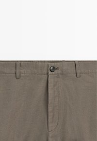 Brązowe spodnie khaki z zapinanym na guziki paskiem w talii, szlufkami na pasek i zamkiem błyskawicznym z przodu, widoczne od talii w górę na białym tle.