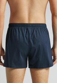 Granatowe kąpielówki z elastycznym pasem, o gładkiej fakturze i krótkiej długości, pokazane od tyłu z nagim tułowiem.