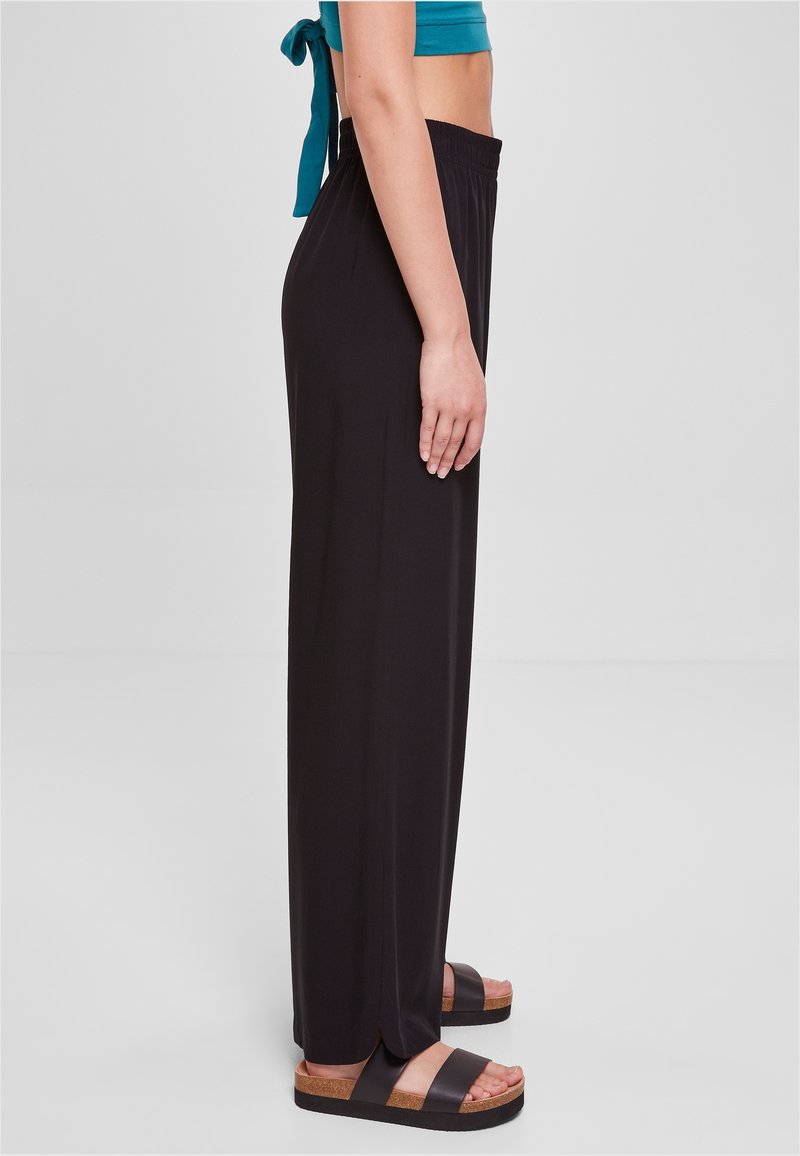 Femme portant un pantalon noir taille haute à jambes larges, un crop top bleu sarcelle et des sandales plateformes noires, debout de profil sur un fond gris.