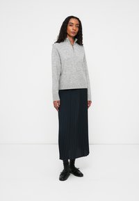 Cárdigan gris con cierre, hecho de un suave tejido de punto, combinado con una larga falda de chiffon azul oscuro. La modelo está de pie frente a un fondo blanco liso.