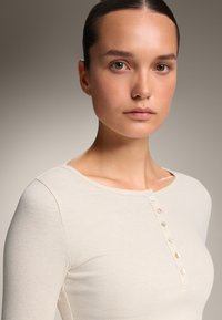 Rippenshirt mit langen Ärmeln in hellem Beige, mit Rundhalsausschnitt und einer Knopfleiste mit mehrfarbigen Knöpfen an der Vorderseite.