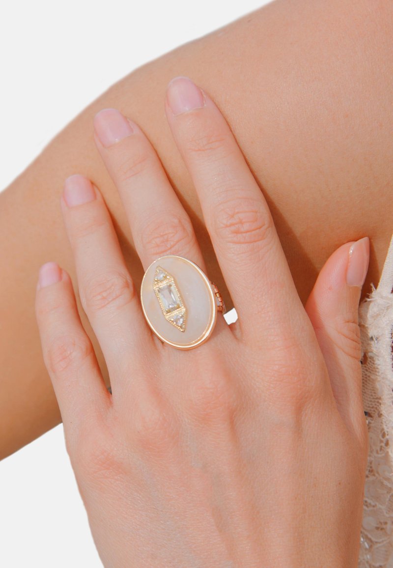 Gold-tone ring with an oval white stone, featuring a central rectangular gem and small accent stones along the edges. Smooth texture.