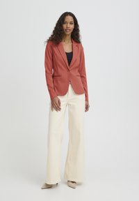 Blazer couleur rouille à coupe ajustée, col cranté, fermeture à un bouton et deux poches avant, associé à un pantalon large couleur crème.