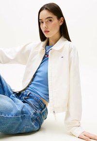 Witte zip-up jas met een kraag, gedragen over een lichtblauw topje. Het model zit in ontspannen blauwe jeans en toont een casual gelaagde stijl.