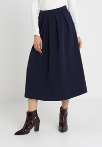 Jupe midi plissée marine en tissu tissé avec une taille haute. Associée à des bottes à talons vernies bordeaux et un haut blanc à manches longues.