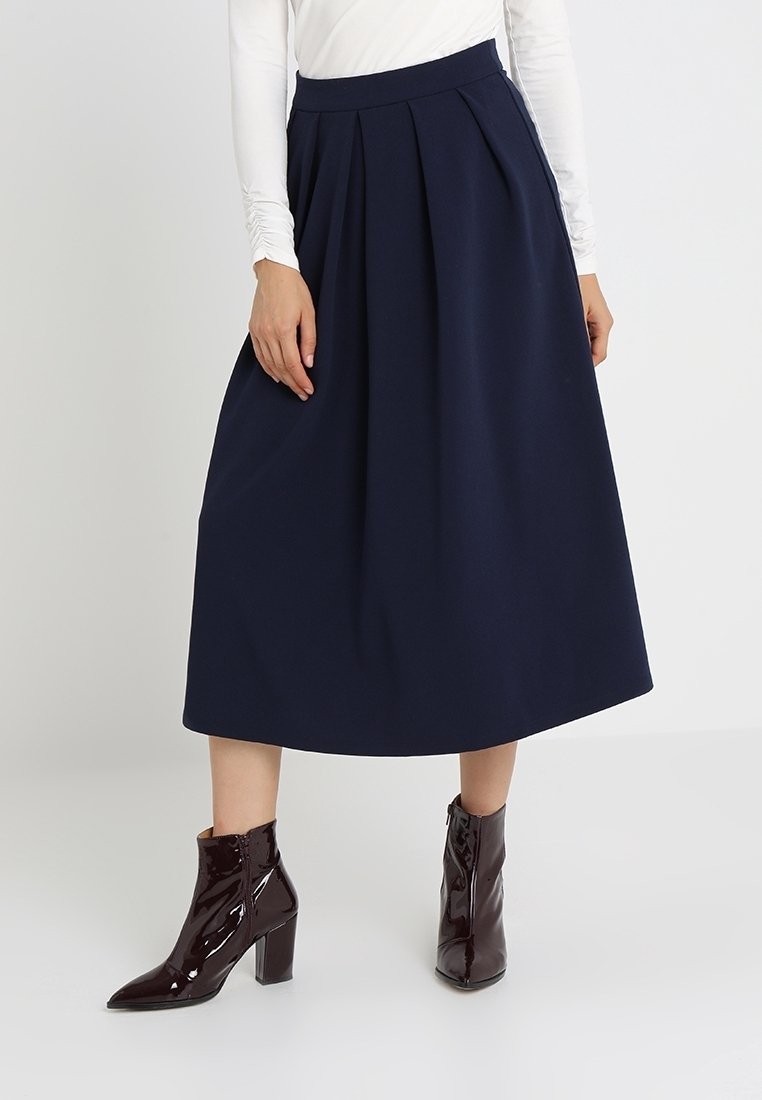 Jupe midi plissée marine en tissu tissé avec une taille haute. Associée à des bottes à talons vernies bordeaux et un haut blanc à manches longues.