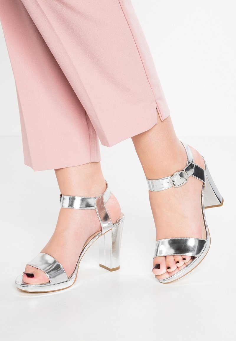 Pieds portant des sandales à talons hauts métalliques argentées avec des brides à la cheville, associées à un pantalon court rose clair sur fond blanc.