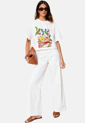 White cotton t-shirt featuring colorful fruit and geometric print, paired with wide-leg white pants and brown strappy sandals, holding a woven bag.