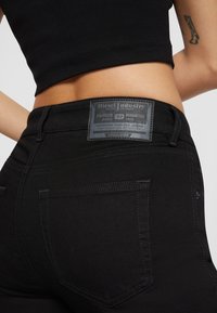 Közeli kép egy személyről, aki fekete, magas derekú farmert visel Diesel Industry folttal és egy fekete crop topot, miközben a karján tetoválás látható.