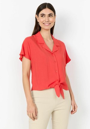 Femme souriante aux cheveux bruns et raides portant un chemisier corail à manches courtes boutonné et noué à la taille, associée à un pantalon beige, debout devant un mur blanc.